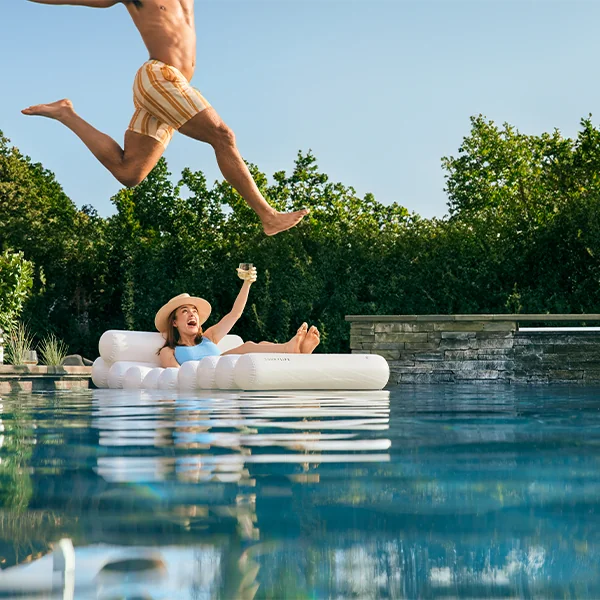 Man jumping into a pool. A woman lounging on a floatie with a glass of Sauvignon Blanc