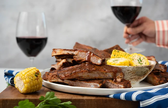Plate of ribs and corn with red wine glasses in the background.